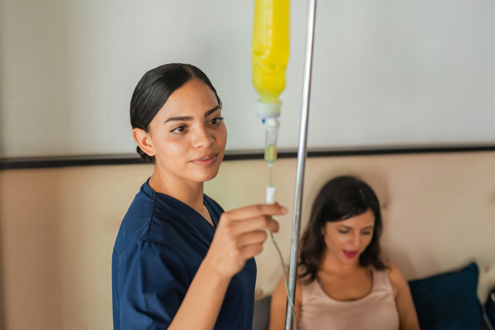 A nurse hangs an IV bag beside a guest sitting on a hotel room bed